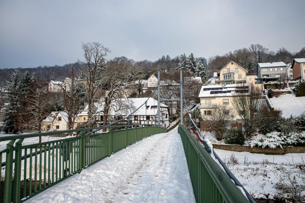 Blick von der anderen Seite der Sieg, über die winterlich verschneite Brücke, auf das Dorf dessen Häuser sich an den Hang schmiegen.